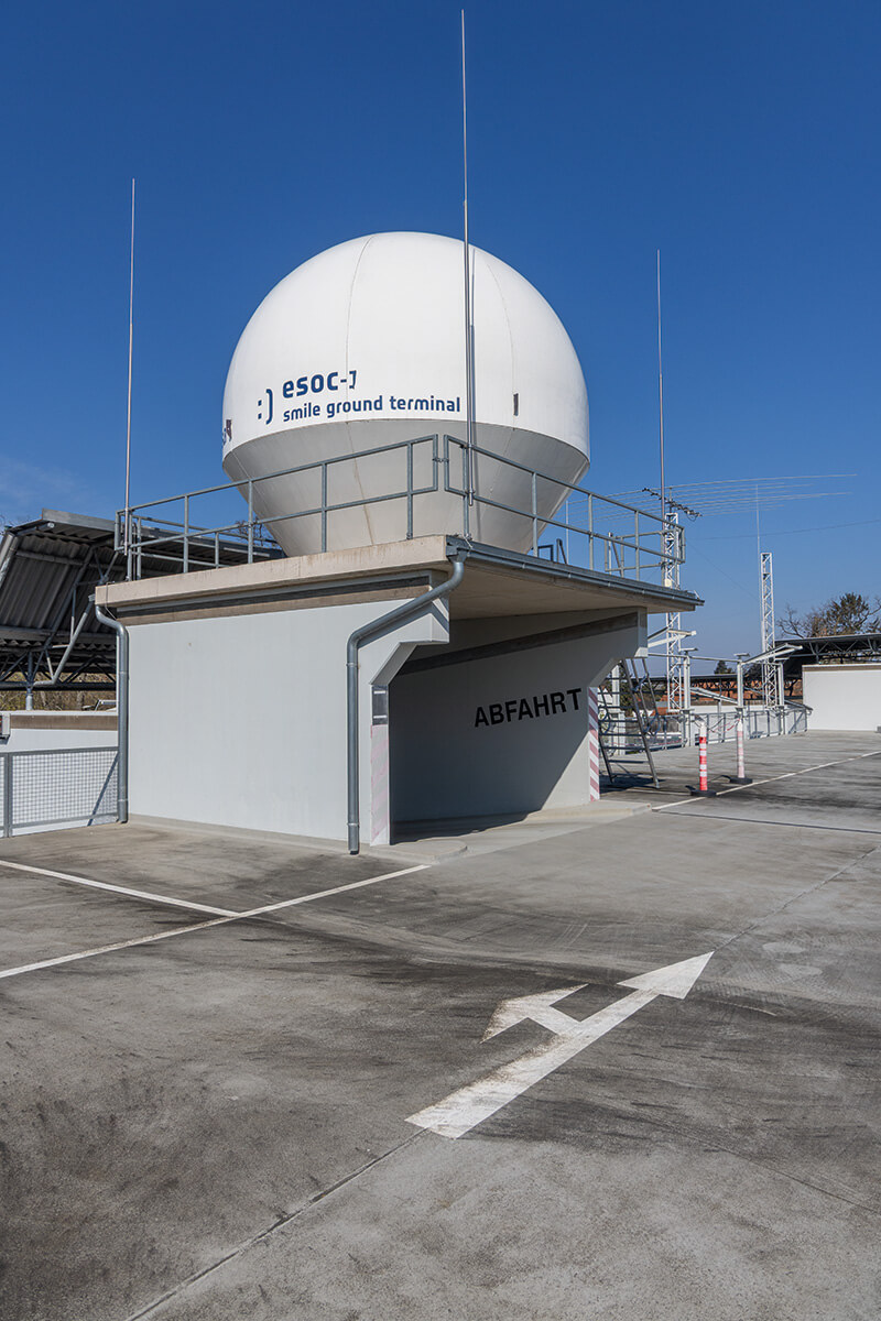 Satellitenbodenstation mit weißer Radarkuppel der „esoc smile ground terminal“ auf einem Flachdach, darunter Einfahrt mit Schriftzug „ABFAHRT“.
