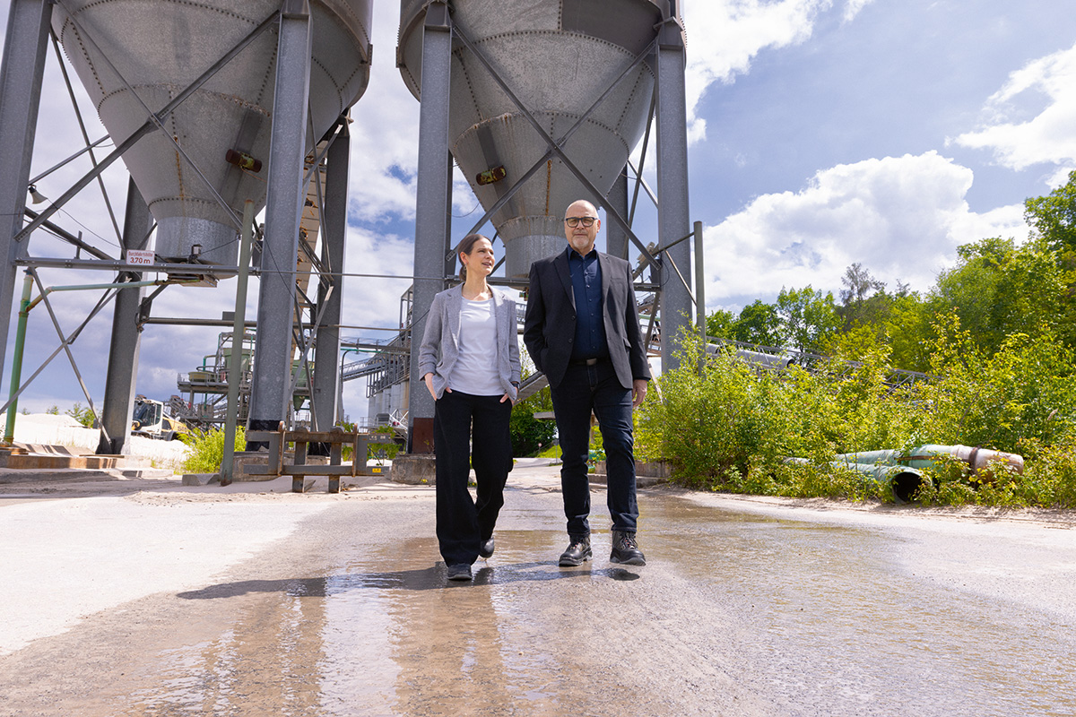 Julia Klinger und Andreas Niedermaier gehen über ein Industriegelände mit großen Silos und technischen Anlagen im Hintergrund.