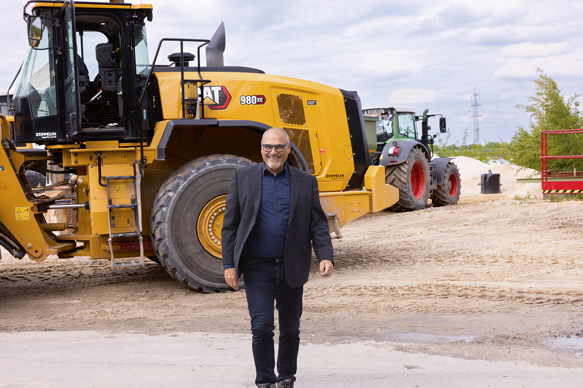 Andreas Niedermaier steht auf einem sandigen Industriegelände vor einem großen gelben Radlader und weiteren Baumaschinen