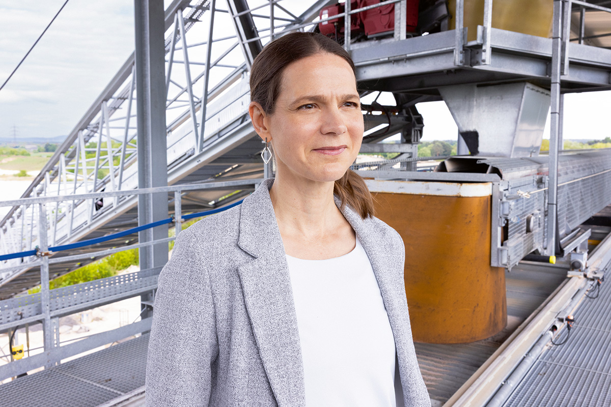 Julia Klinger steht auf einer technischen Anlage mit Metallkonstruktionen und Fördertechnik im Hintergrund.