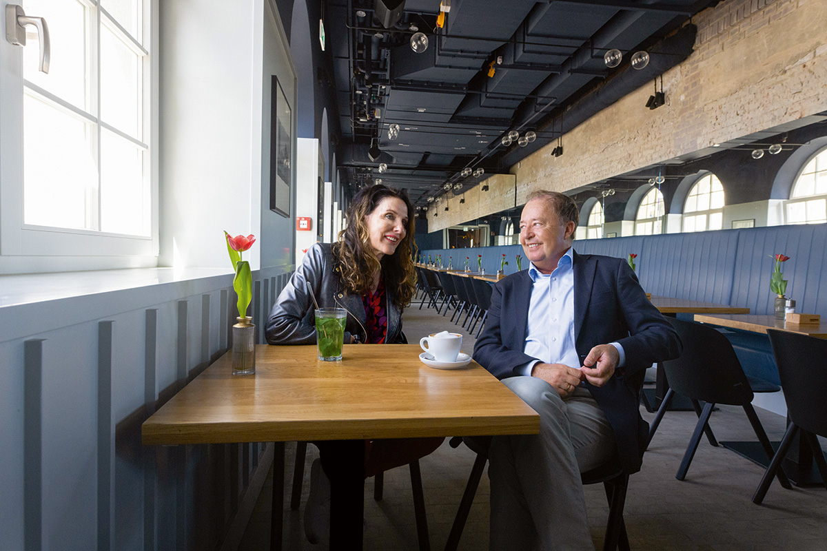 Anja Förster und Thomas Schmidt sitzen in einem hellen Cafébereich im Gespräch. Auf dem Tisch stehen Getränke und eine rote Tulpe am Fenster.