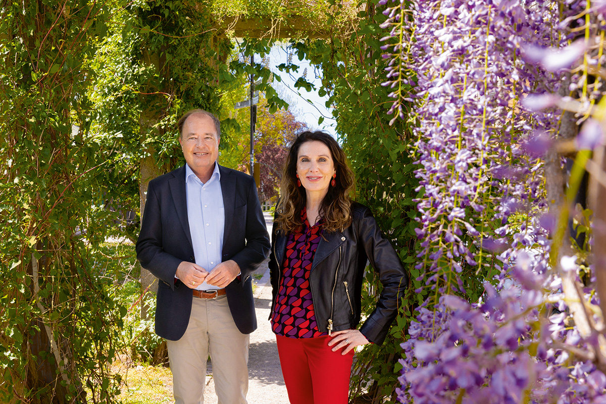 Anja Förster und Thomas Schmidt stehen in einem begrünten Laubengang auf der Mathildenhöhe Darmstadt, eingerahmt von violett blühendem Blauregen.