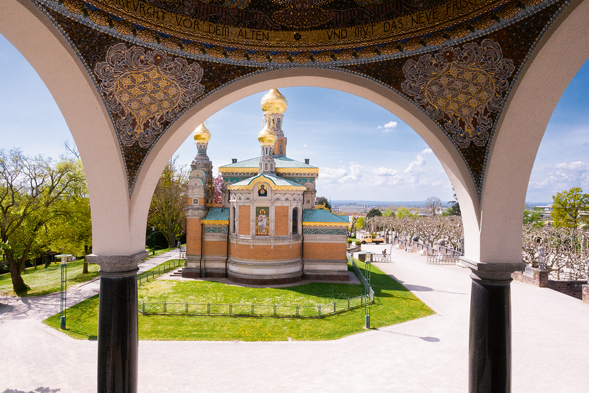 Blick durch einen verzierten Rundbogen auf die Russische Kapelle St. Maria Magdalena auf der Mathildenhöhe Darmstadt. Die Kapelle mit goldenen Zwiebeltürmen steht auf einer grünen Rasenfläche, dahinter öffnet sich der Blick über die Stadt.