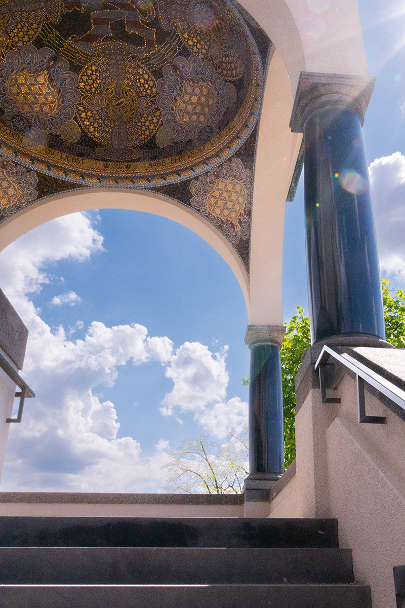 Verzierter Rundbogen mit Mosaikdecke auf der Mathildenhöhe Darmstadt. Der Blick führt von einer Treppe aus zwischen dunklen Säulen hindurch in den hellen Himmel.