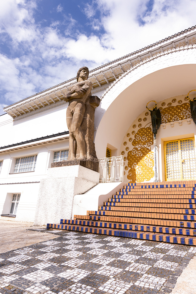 Eingang des Ausstellungsgebäudes auf der Mathildenhöhe Darmstadt mit breiter, blau abgesetzter Treppe, goldverzierter Portalnische und einer monumentalen Steinfigur vor heller Jugendstil-Architektur.